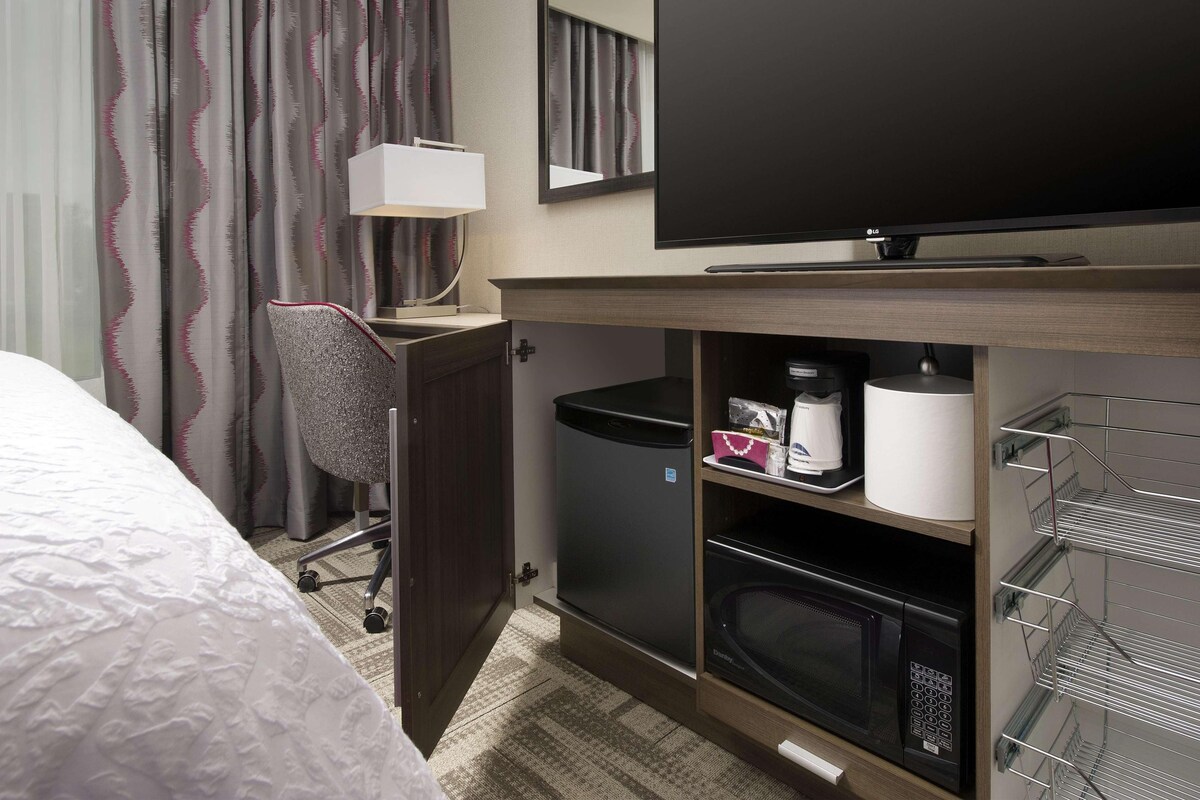 Habitación de hotel con una cama, un escritorio con silla, un televisor, un microondas, un cubo de basura y un estante con artículos de tocador.