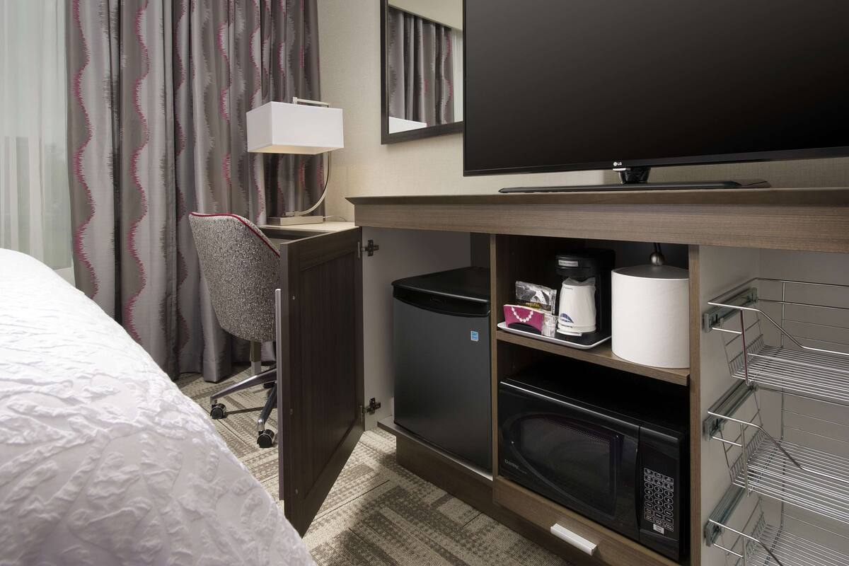 Habitació d'hotel amb un llit, una taula de treball amb una cadira, una televisió, un microones, un gresol i una estantària amb productes de tocador.