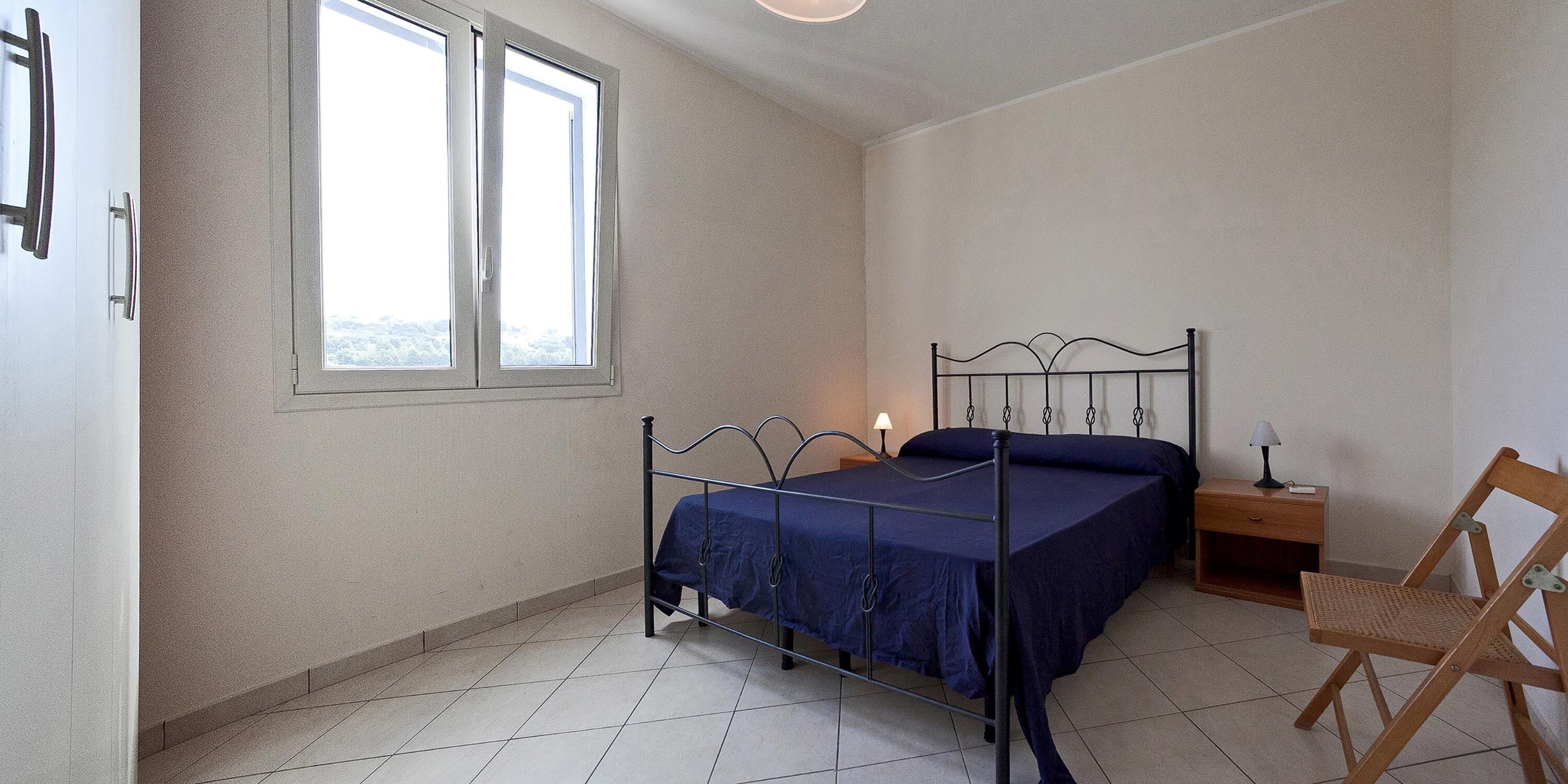 Ein Schlafzimmer mit einem Metallbettgestell, einem Holzstuhl, einem Nachttisch mit Lampe und einem Fenster mit Blick auf Bäume.