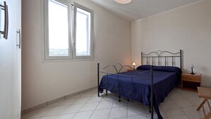 A bedroom with a metal bed frame, a wooden chair, a nightstand with a lamp, and a window with a view of trees. - Casa del Sole Trilo A (Castrignano del Capo)