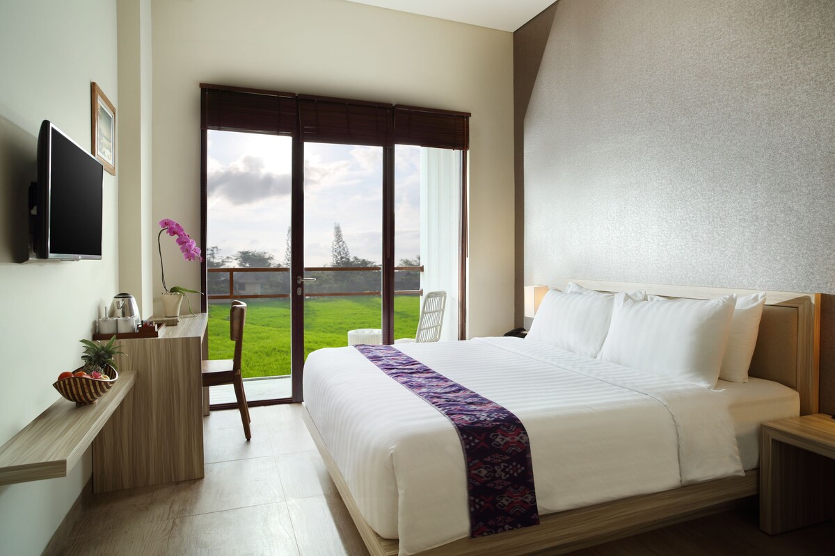 A hotel room with a bed, a desk, a chair, a TV, and a balcony with a view of the sky and greenery.