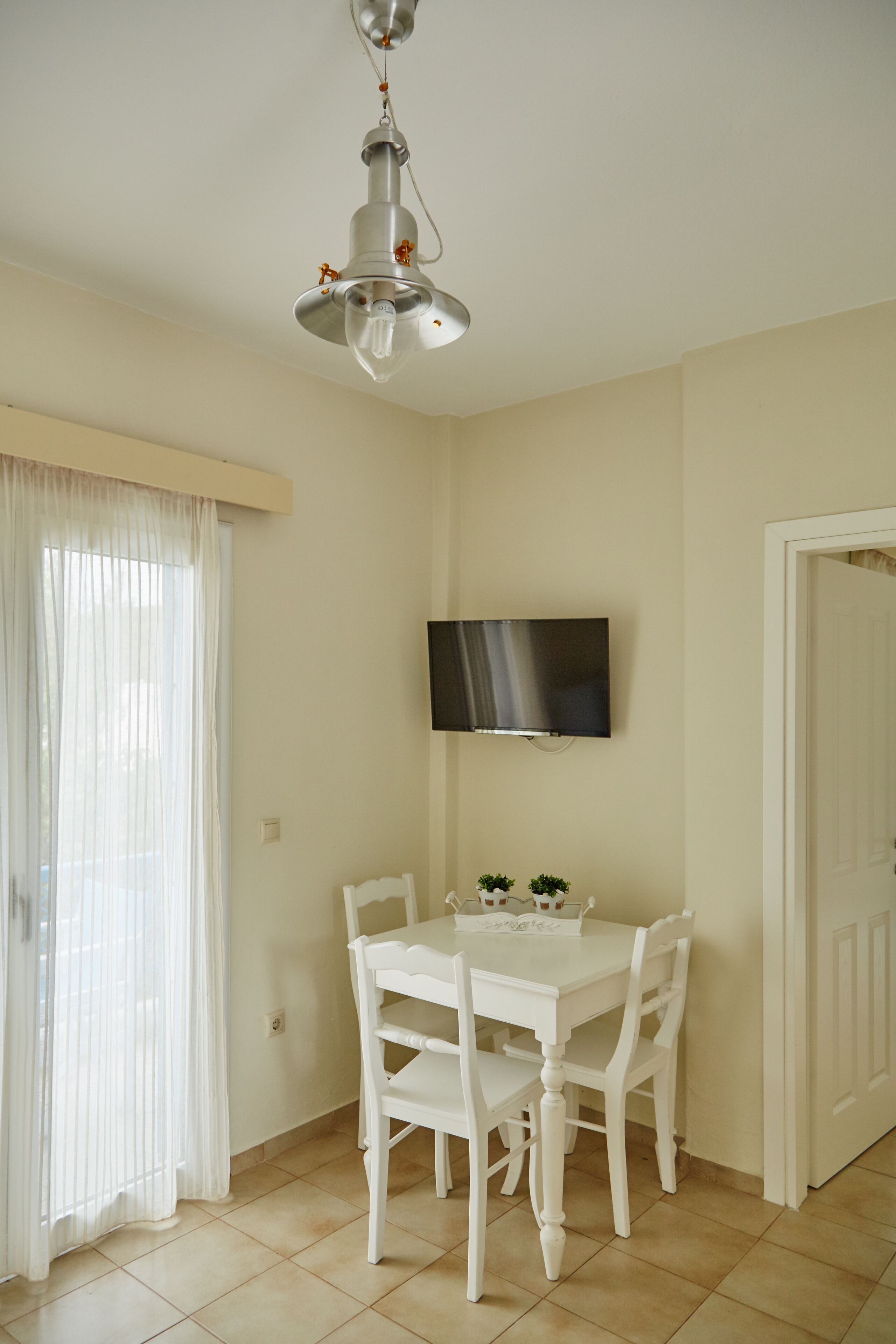 Un coin repas avec une table et des chaises blanches, un luminaire au plafond, un téléviseur à écran plat fixé au mur et une porte sur le côté droit.