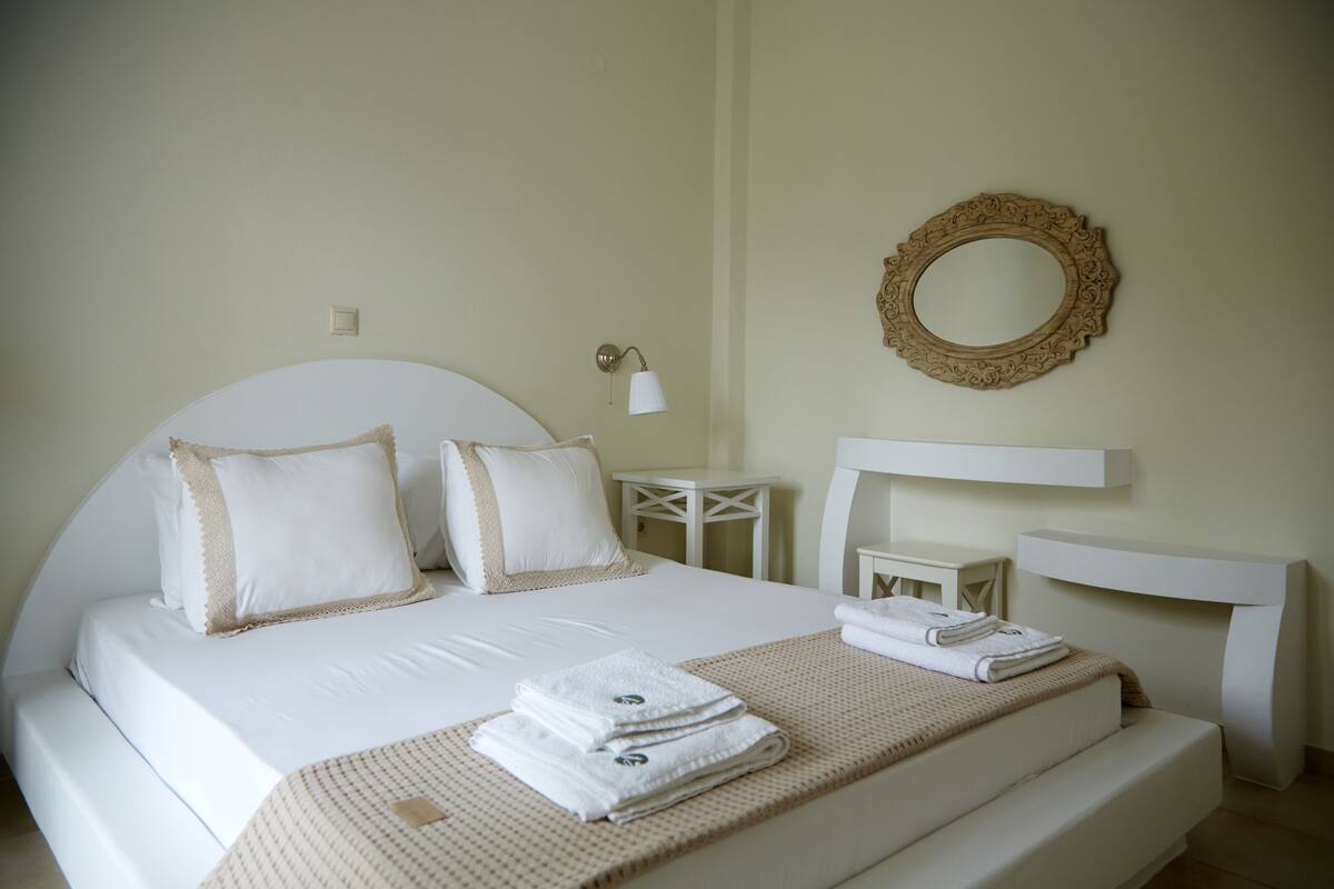 A neatly made bed with white linens and a beige throw blanket, a bedside table with a lamp, and a wall-mounted mirror.