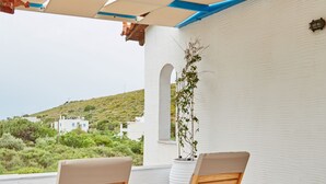 A covered outdoor seating area with two wooden chairs and a small table, overlooking a hillside with white buildings. - Aelia Apartments (Andros)