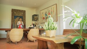 A room with wicker chairs, a wooden table, a potted plant, a painting, and a person standing by a counter. - Bumi Muwa Ubud (Ubud)