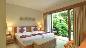 A hotel room with two beds, a wooden headboard, a nightstand with a lamp, a wicker bench, and a view of greenery through the window. - Bumi Muwa Ubud (Ubud)