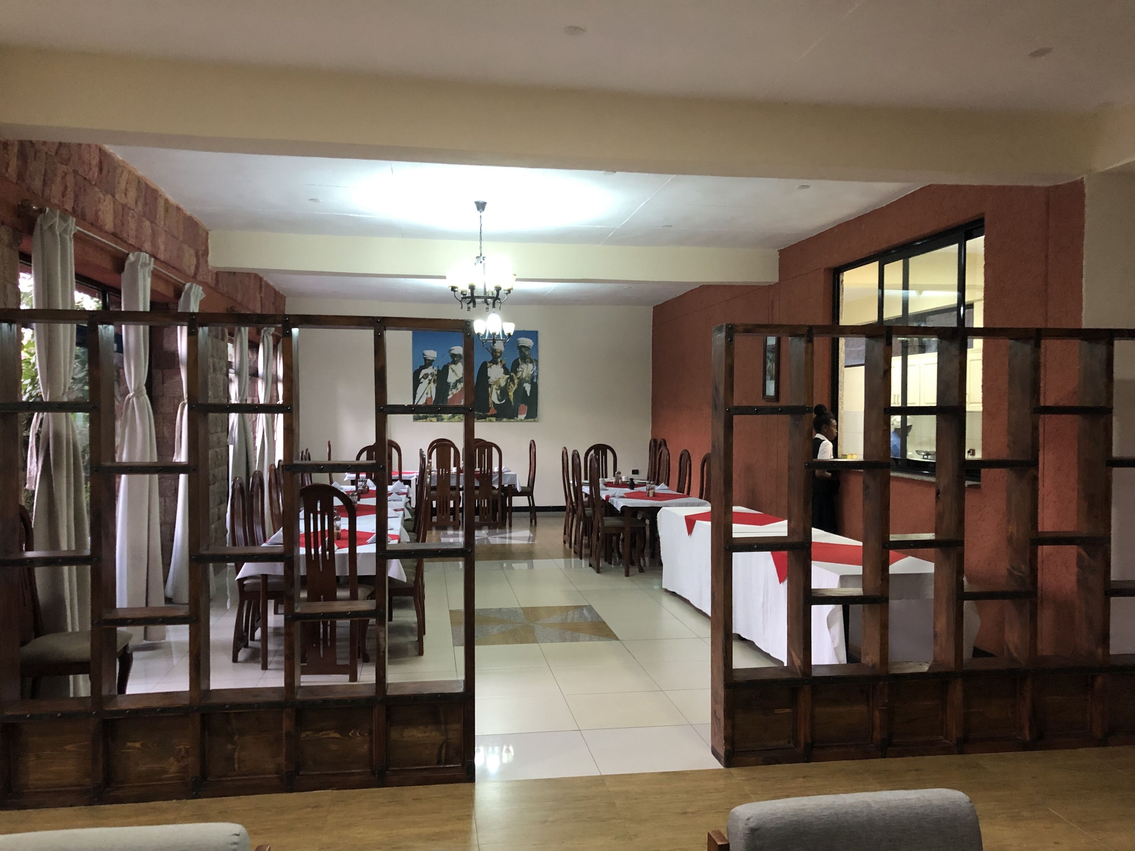 Comedor con mesas dispuestas para varias personas, sillas de madera, una araña y un cuadro grande en la pared.