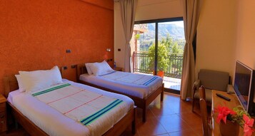 Habitación de hotel con dos camas, un balcón con vistas, un escritorio y una silla, y un televisor.