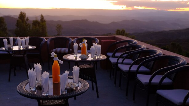 Mesas y sillas para comer al aire libre, dispuestas en una terraza con vistas a montañas distantes.