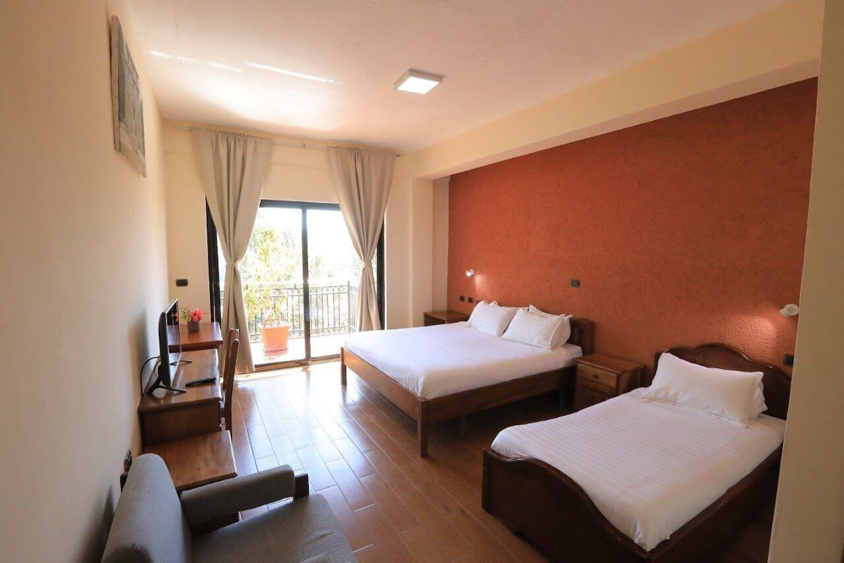A hotel room with two beds, a desk, a chair, and a balcony with curtains.