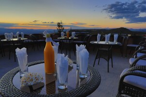 Un área de comedor en la azotea con mesas preparadas para dos, cada una con una botella, vasos y servilletas.