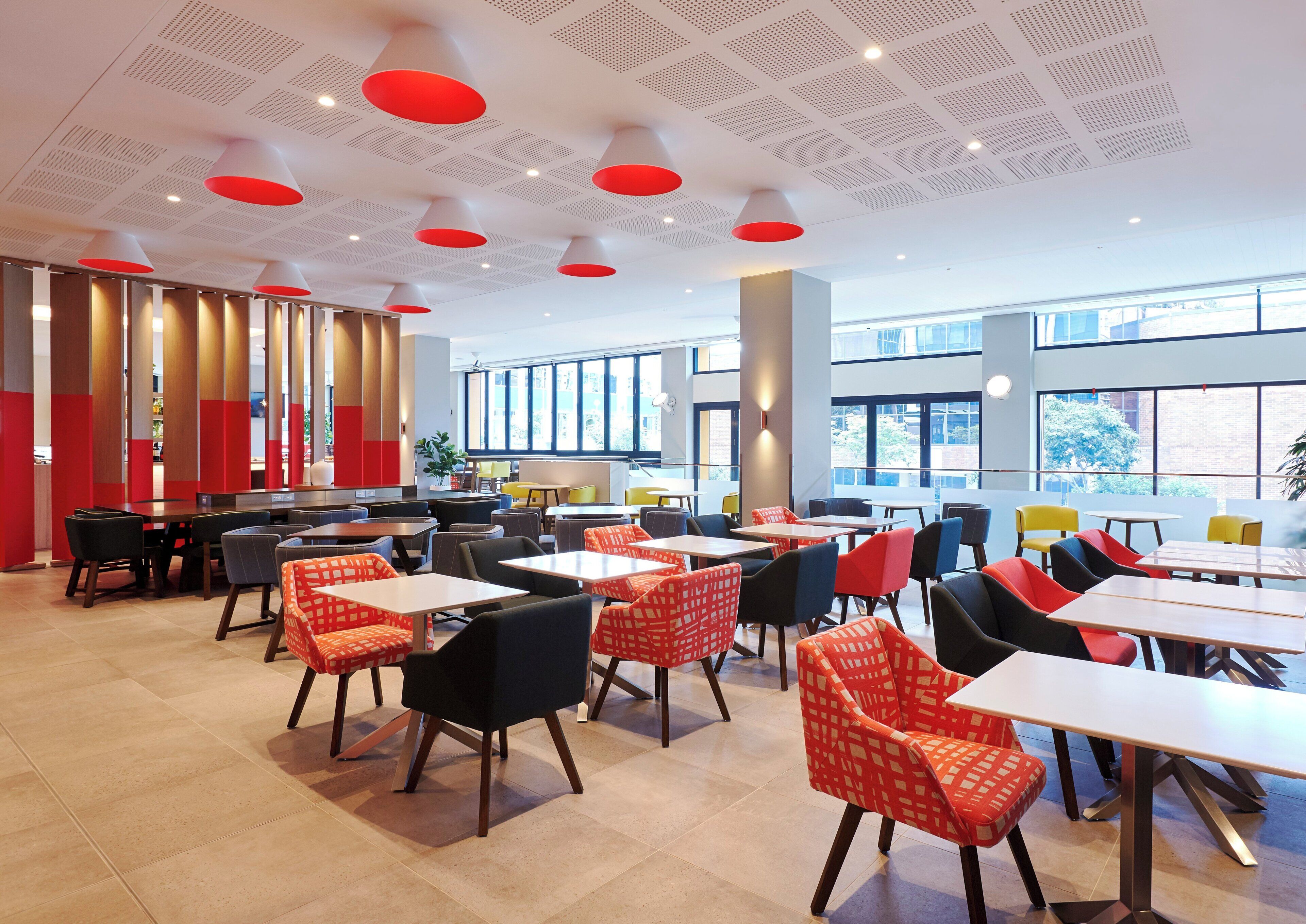 A modern dining area with multiple tables and chairs, large windows, and a ceiling with red accents.