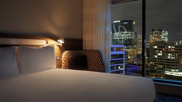 Hotelkamer met een groot bed, nachtkastje en uitzicht op een stadsbeeld 's nachts.