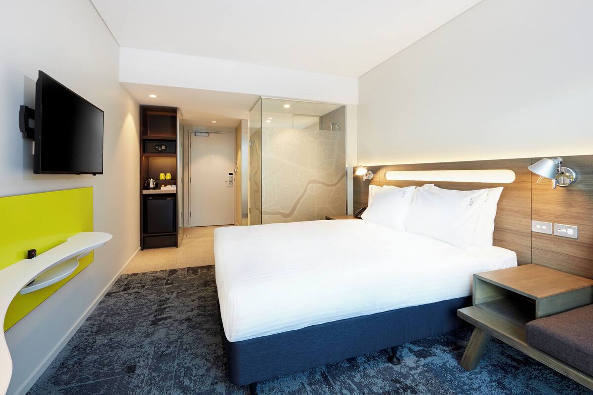 Une chambre d’hôtel moderne dotée d’un grand lit, d’un téléviseur à écran plat fixé au mur, d’une petite kitchenette et d’une cabine de douche en verre.