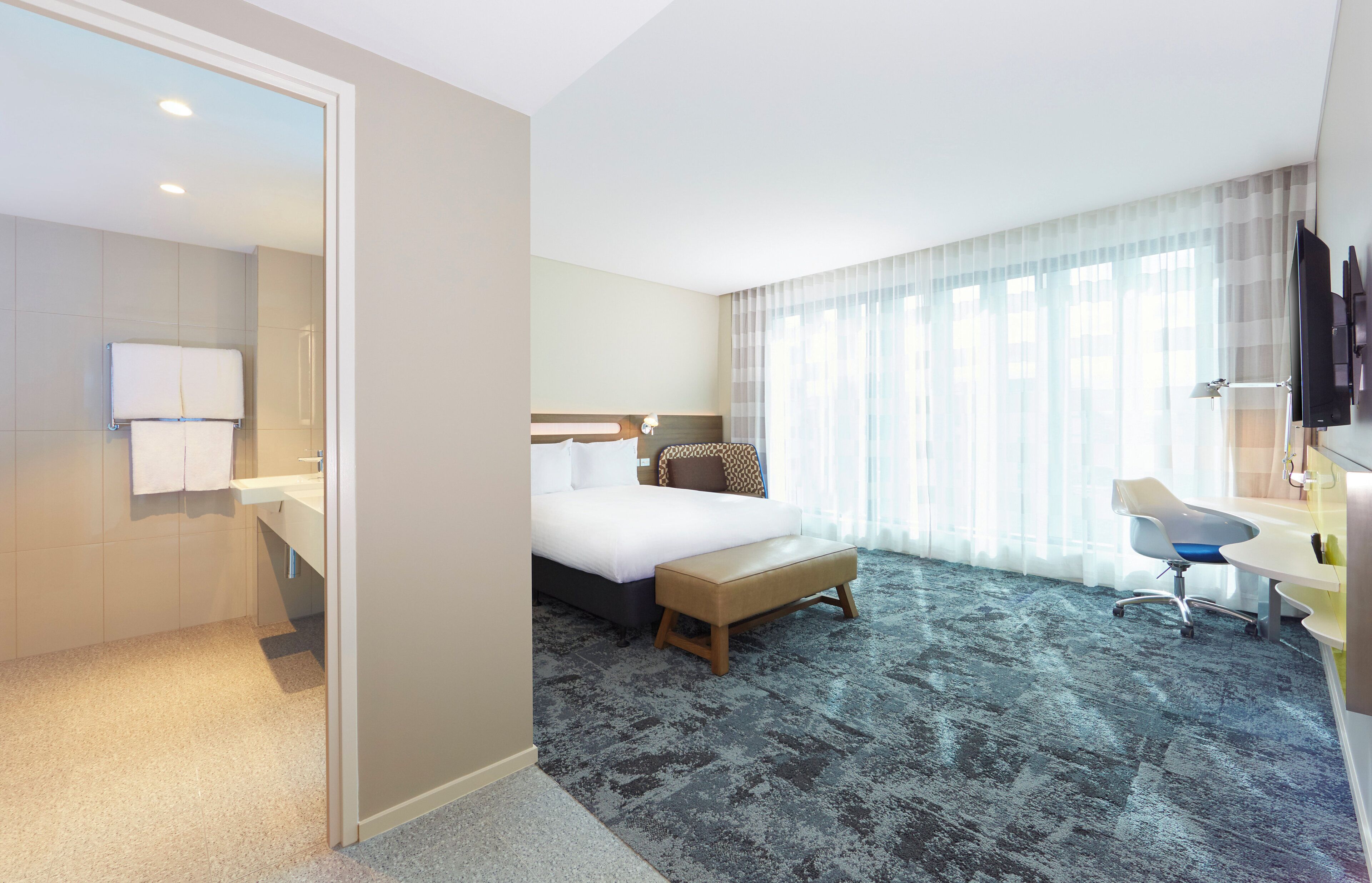 A hotel room with a bed, bedside table, chair, desk, and a large window with curtains.