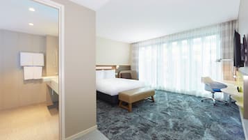 A hotel room with a bed, bedside table, chair, desk, and a large window with curtains.