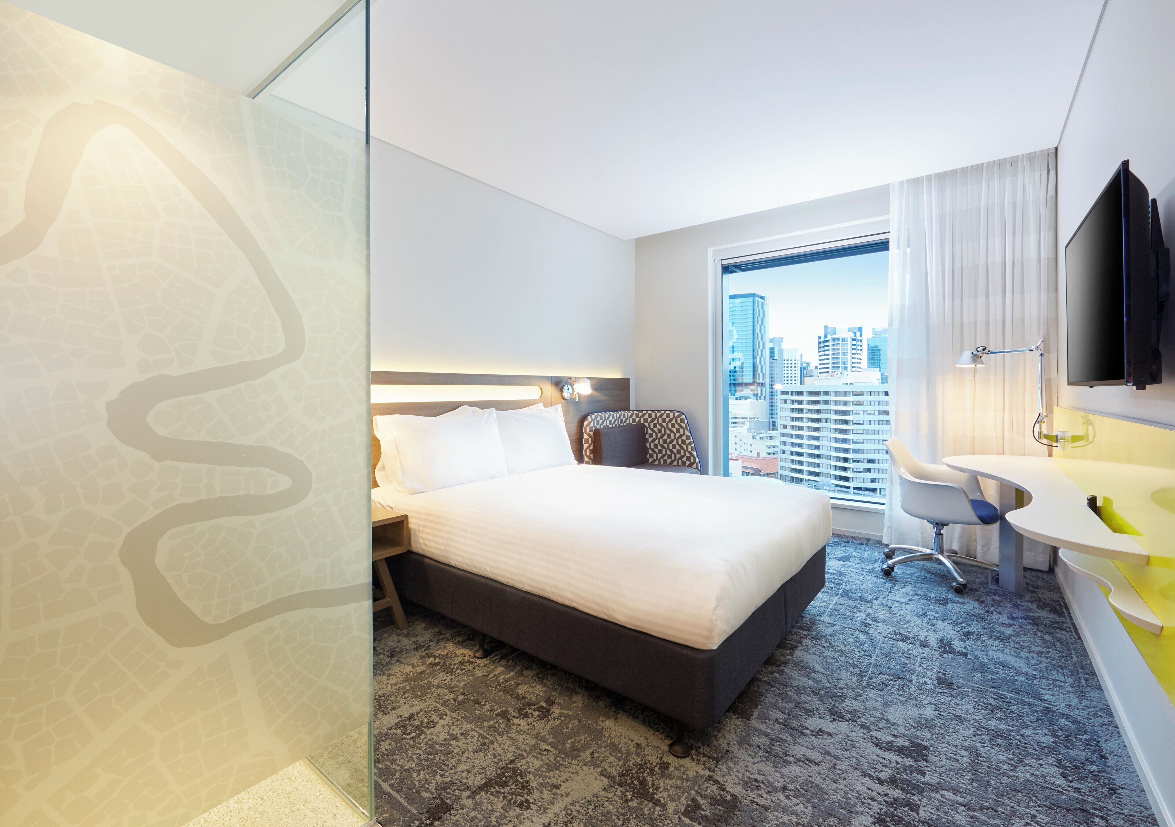 A hotel room with a bed, a desk, a chair, a window with a city view, and a bathroom with a sink and mirror.