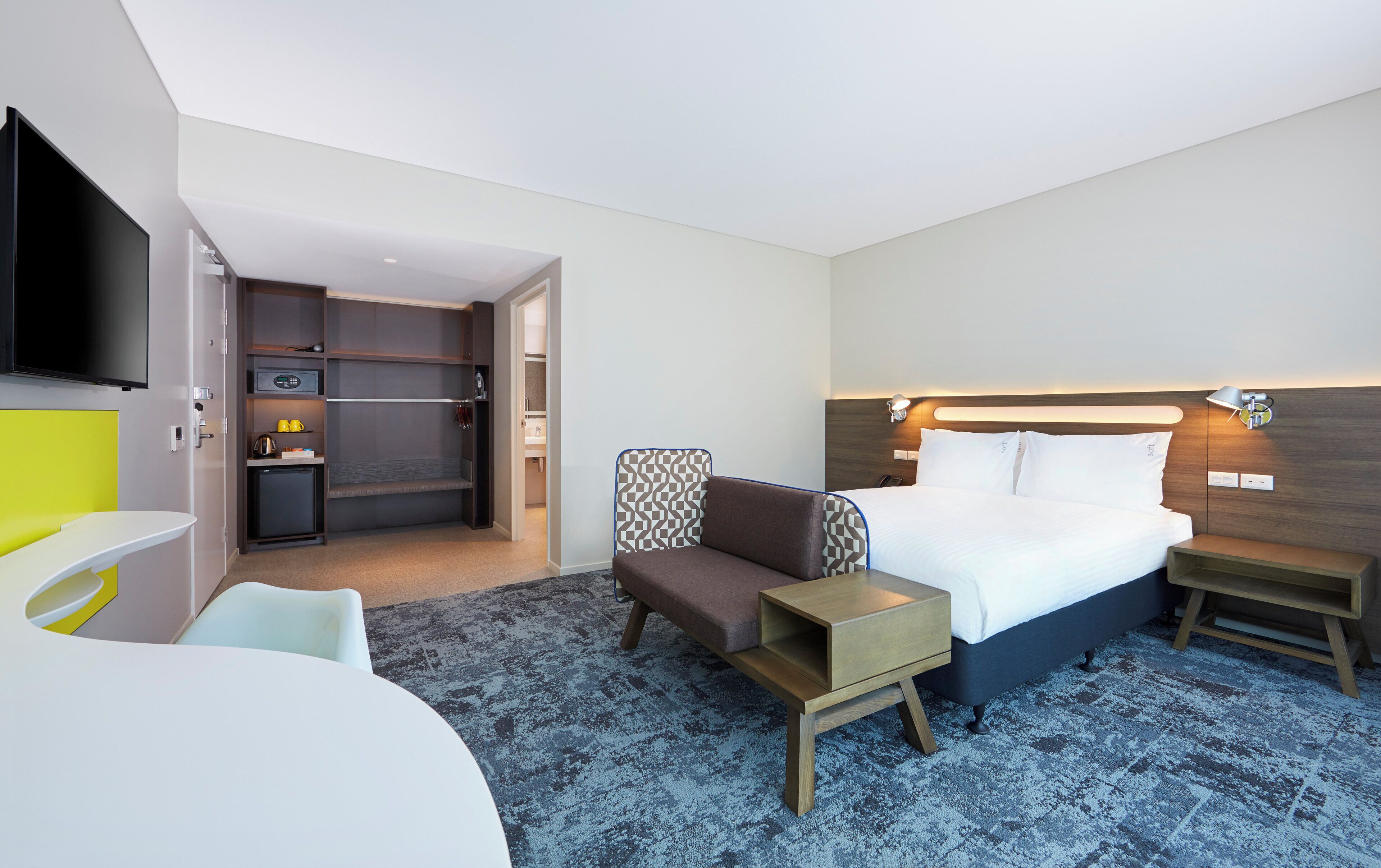A hotel room with a bed, bedside table, chair, and a small desk with a chair. There is a kitchenette in the background.