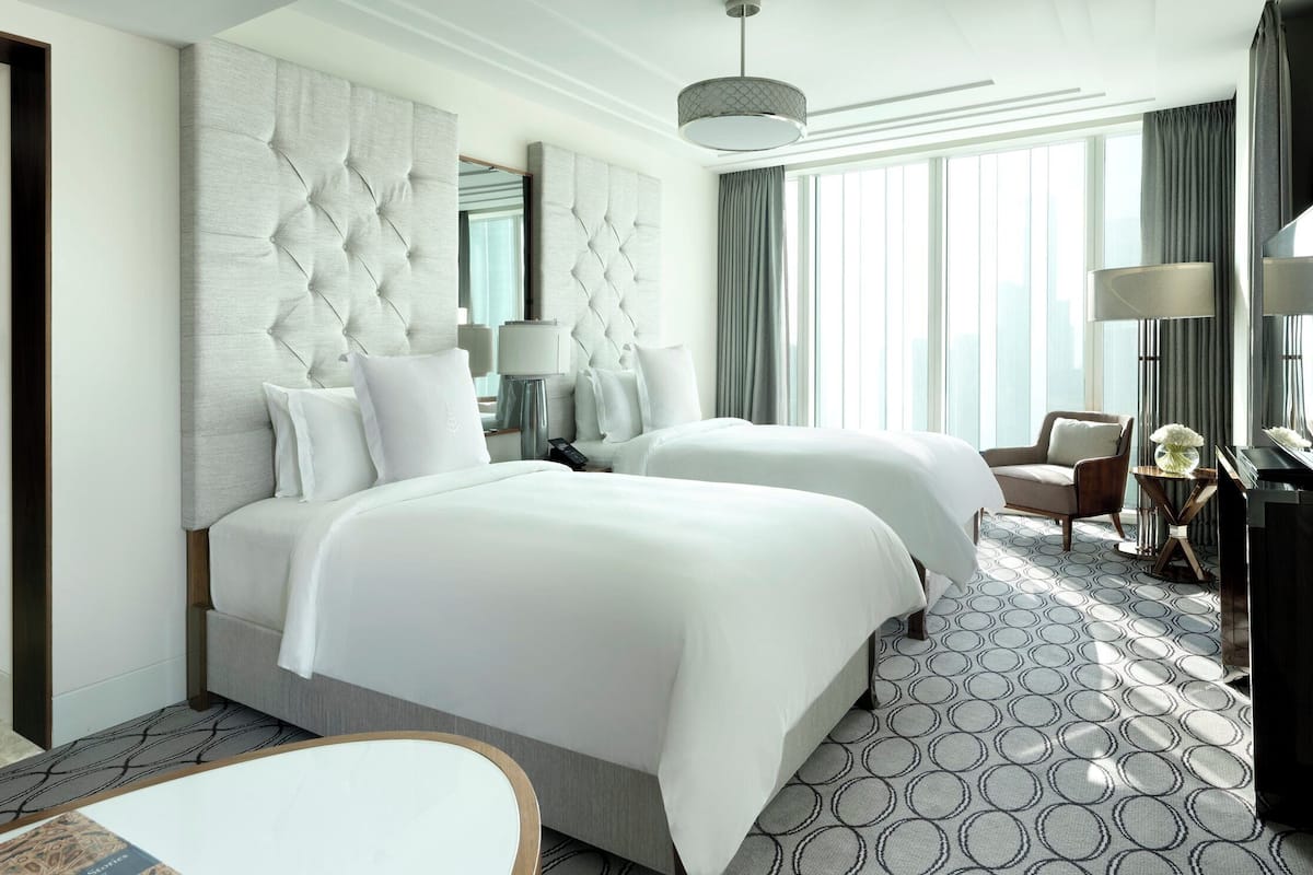 Ein Hotelzimmer mit zwei Betten, einem großen, gepolsterten Kopfteil, einem Schreibtisch mit Lampe, einem Stuhl und Blick auf die Stadt durch große Fenster.
