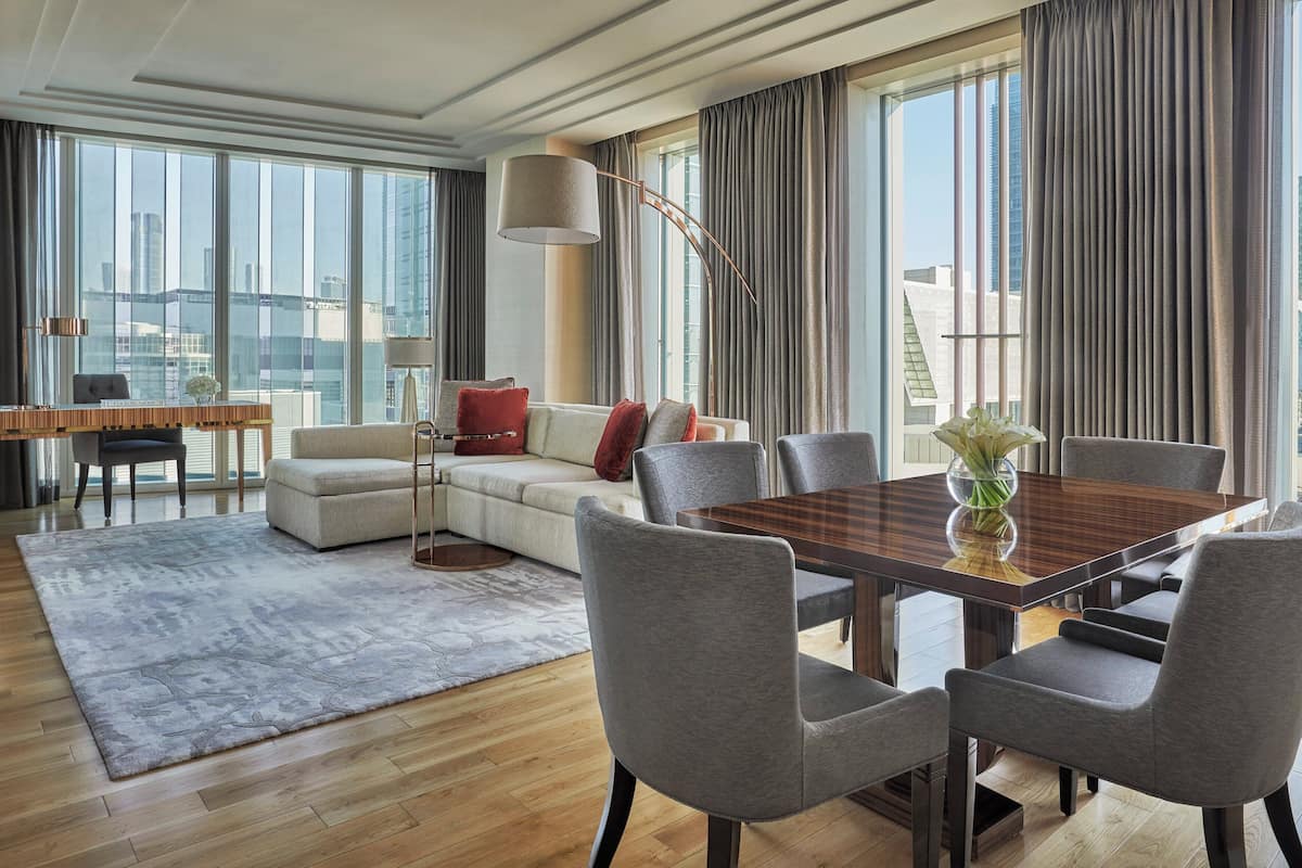 Ein modernes Wohnzimmer mit einem großen Esstisch, grauen Polsterstühlen, einer Couch mit roten Kissen und Blick auf die Stadt durch große Fenster.