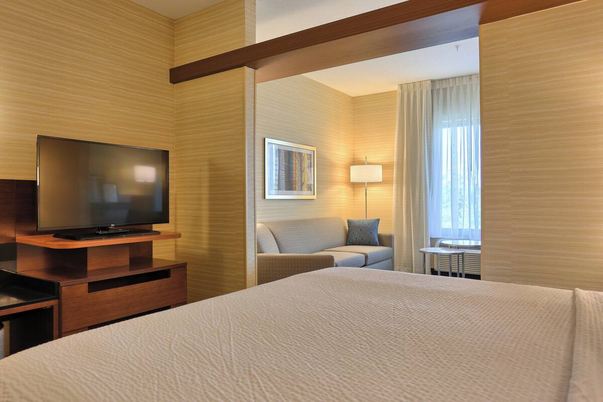 A hotel room with a large bed, a flat-screen TV on a wooden stand, a sofa with a pillow, a small table, and a window with curtains.
