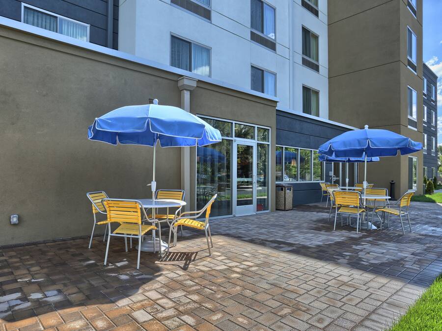 Een terras buiten met twee tafels en stoelen, elk onder een blauwe parasol, en een glazen deur die naar een gebouw leidt.