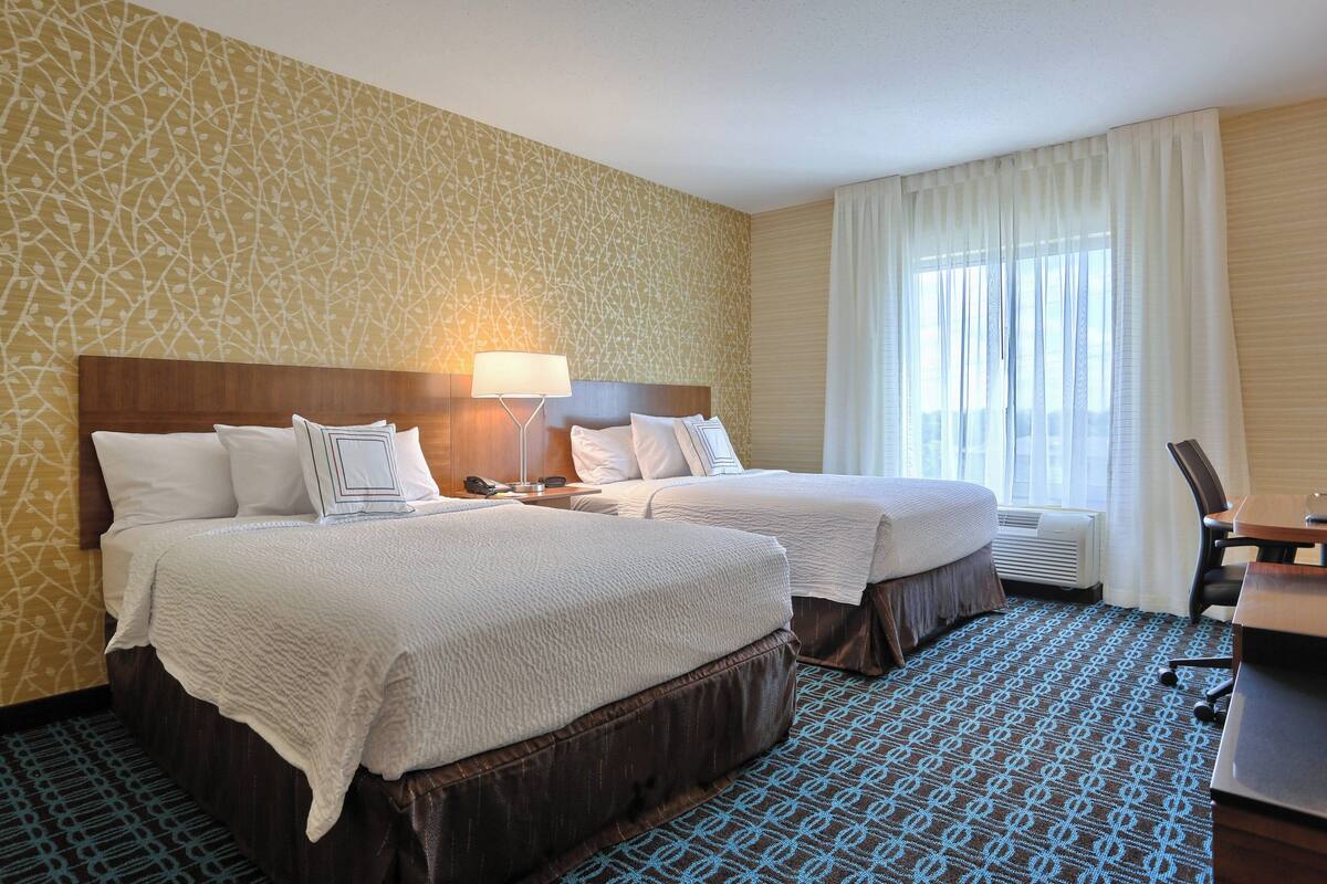 A hotel room with two beds, a desk, a chair, a lamp, and a window with curtains.