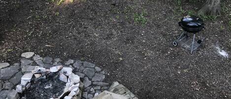 Un espacio al aire libre rodeado de árboles con un hogar de piedra y una parrilla portátil.