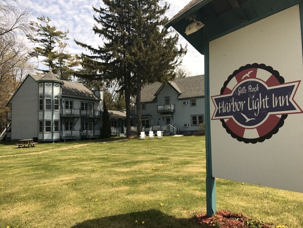 A sign for Gills Rock Harbor Light Inn with a view of a large, multi-story building with a balcony and a lawn with white chairs.