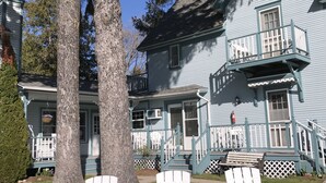 A two-story house with a balcony, white chairs in front, and a large tree.