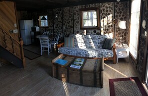 A cozy cabin interior with a stone wall, wooden furniture, a sofa with a nautical print, a dining table, and a kitchen area with a refrigerator and microwave.