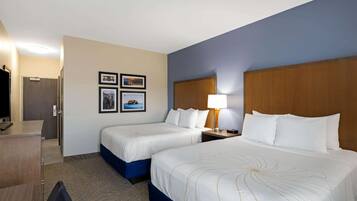 A hotel room with two beds, a desk, a chair, and a TV. There are framed pictures on the wall and a lamp on the nightstand.