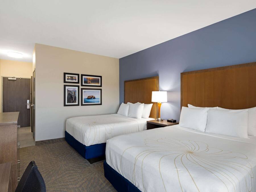 A hotel room with two beds, a desk, a chair, and a TV. There are framed pictures on the wall and a lamp on the nightstand.