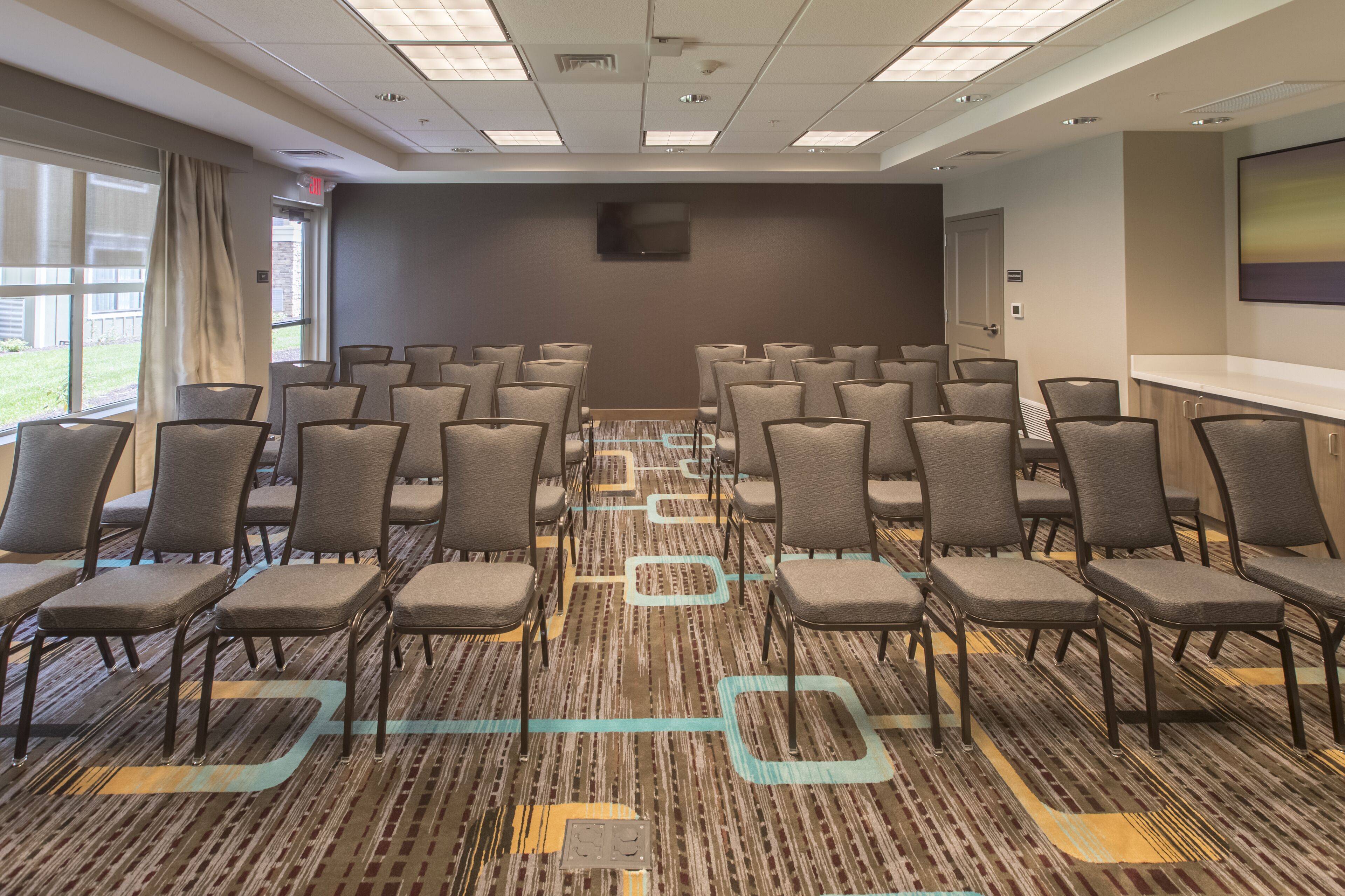 A conference room with rows of chairs, a carpet with geometric patterns, a flat-screen TV on the wall, and large windows with curtains.