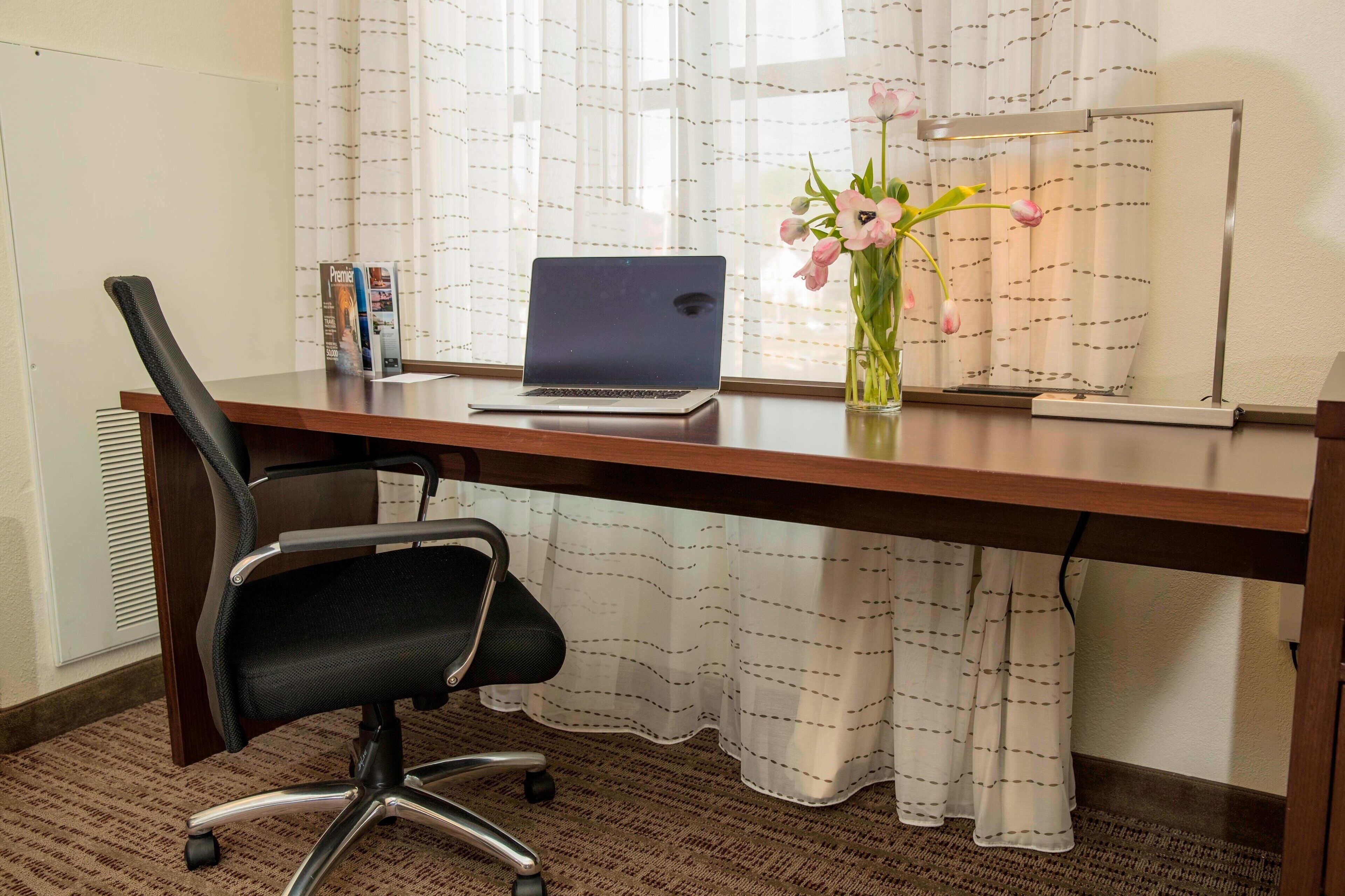 Un bureau en bois avec un ordinateur portable, une chaise de bureau noire, un vase avec des fleurs et un lampadaire.