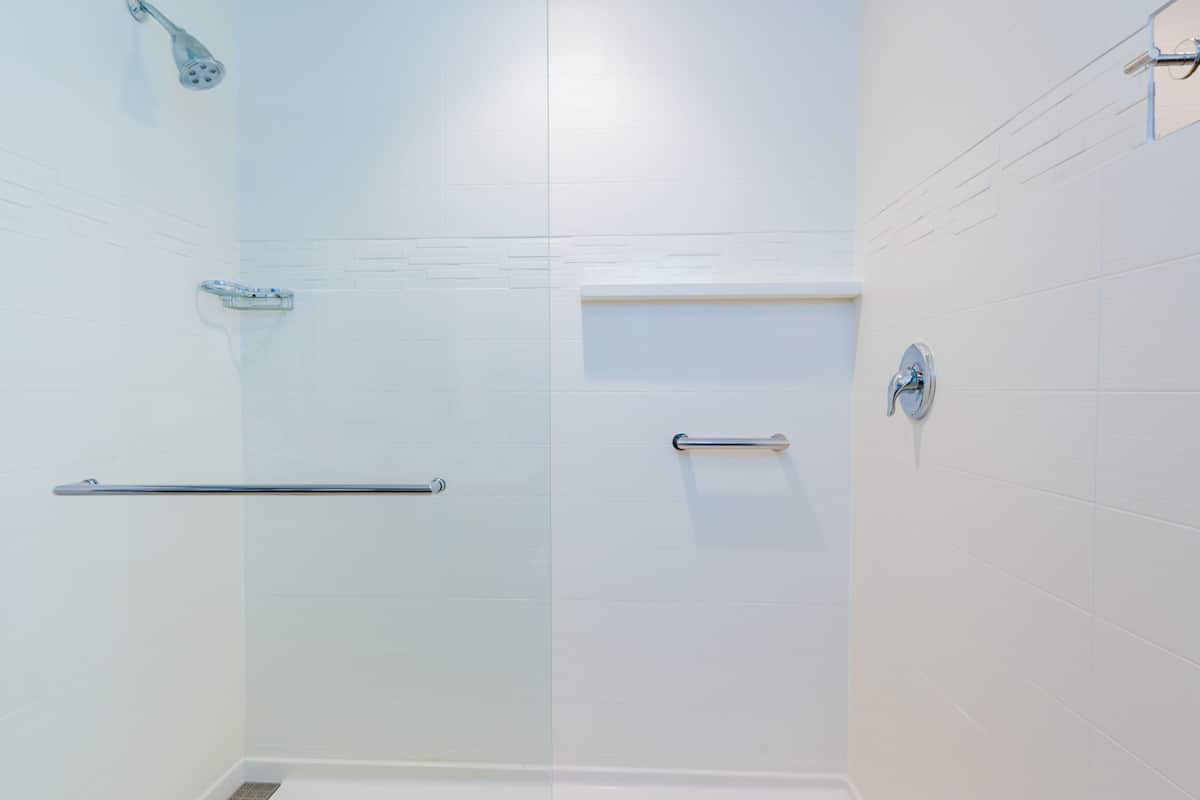 A modern bathroom with white tiles, a glass shower enclosure, a handrail, and a towel rack.