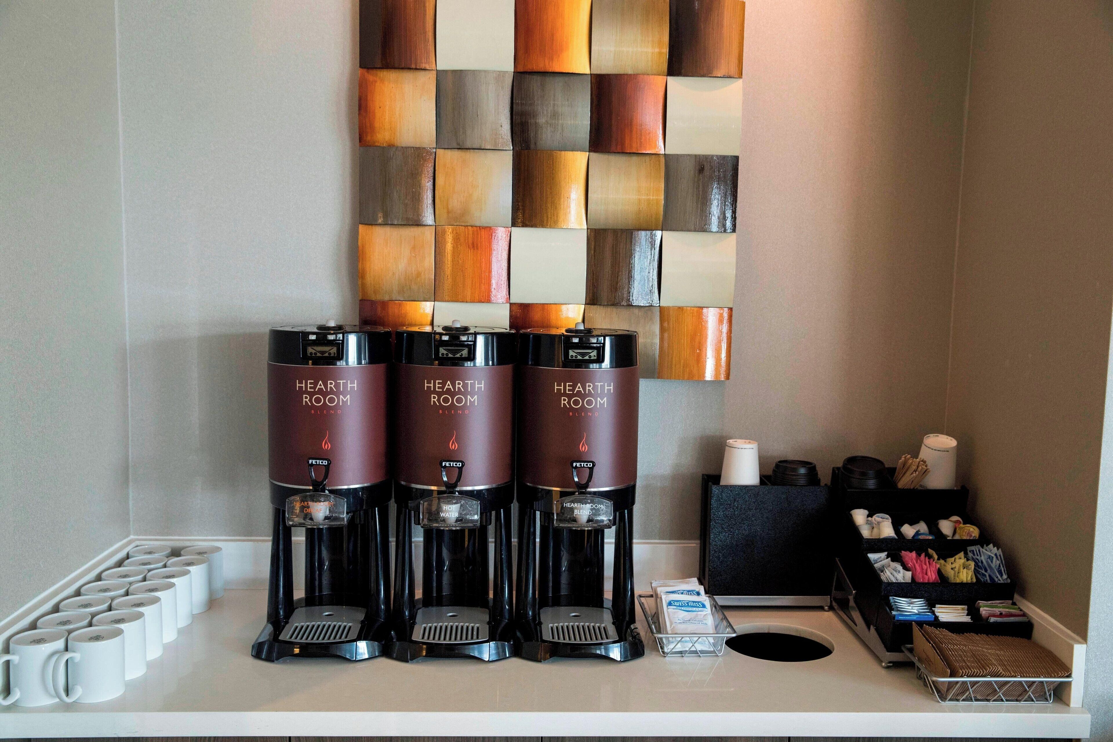 Three coffee machines labeled "HEARTH ROOM" on a counter with disposable cups and a tray of utensils.