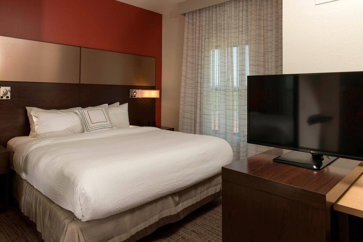 A hotel room with a large bed, a flat-screen TV, a wooden desk, and a window with curtains.