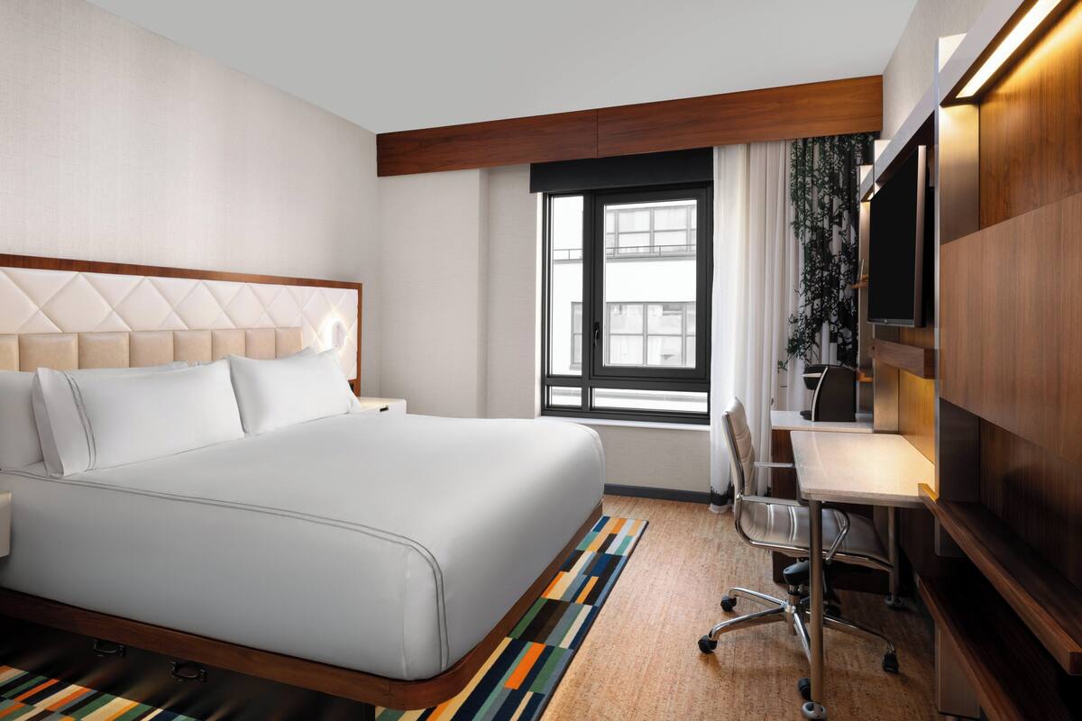 Une chambre d’hôtel avec un grand lit, un bureau, une chaise, une télévision et une fenêtre avec des stores.