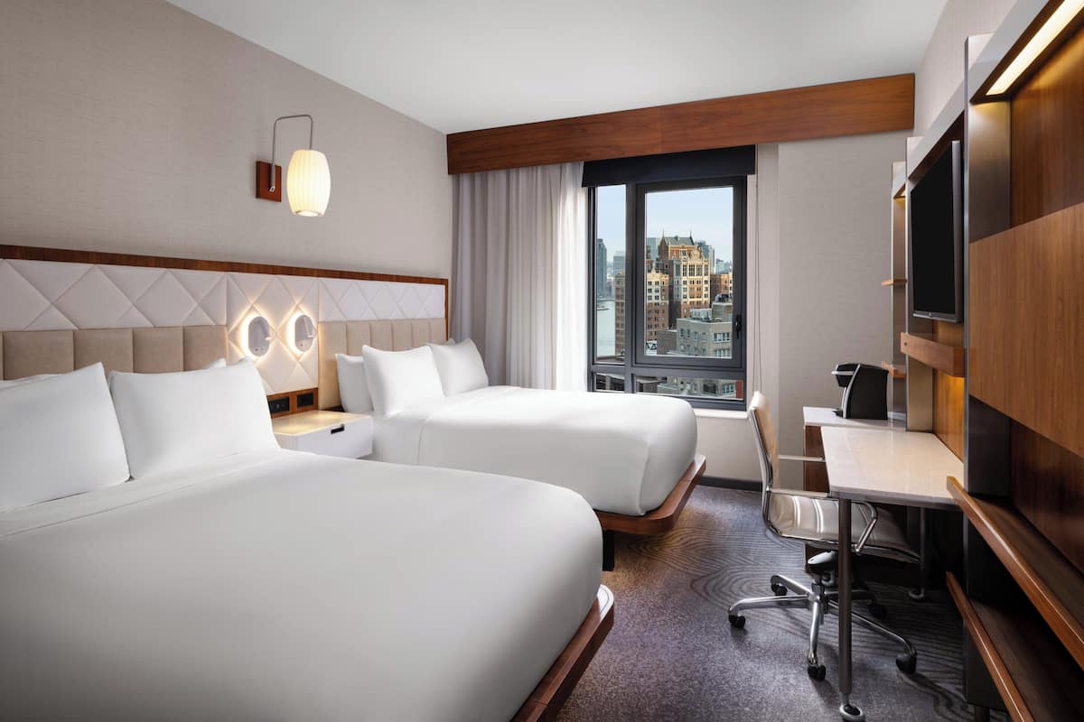 Une chambre d’hôtel avec deux lits, un bureau, une chaise, une télévision et une grande fenêtre donnant sur la ville.