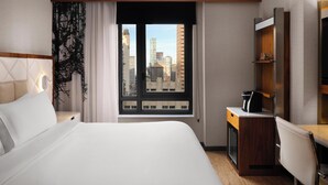Ein Hotelzimmer mit einem Bett, einem Schreibtisch mit Kaffeemaschine, einem Fernseher und einem Fenster mit Blick auf die Stadt.