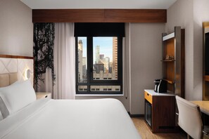 Een hotelkamer met een bed, een bureau met koffiezetapparaat, een televisie en een raam met uitzicht op de stad.