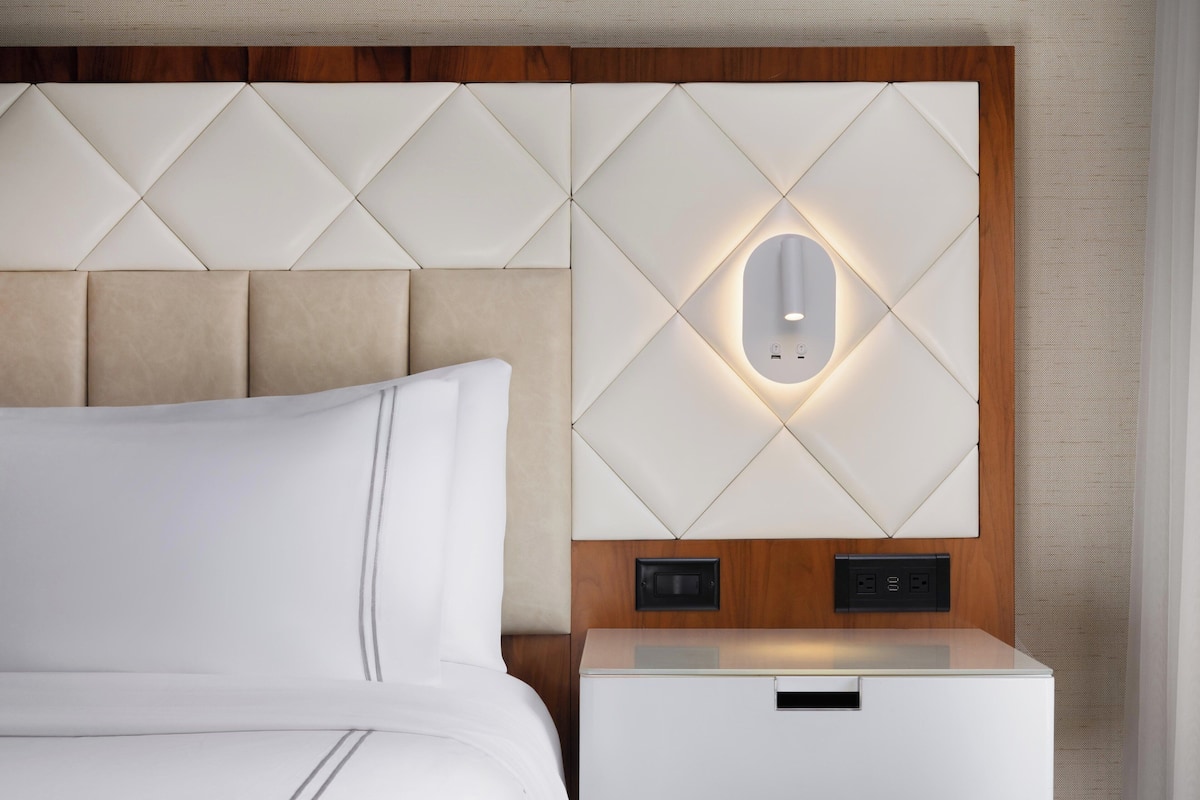 Une chambre d’hôtel moderne dotée d’une tête de lit capitonnée, d’une table de chevet avec une lampe intégrée et d’un luminaire fixé au mur.