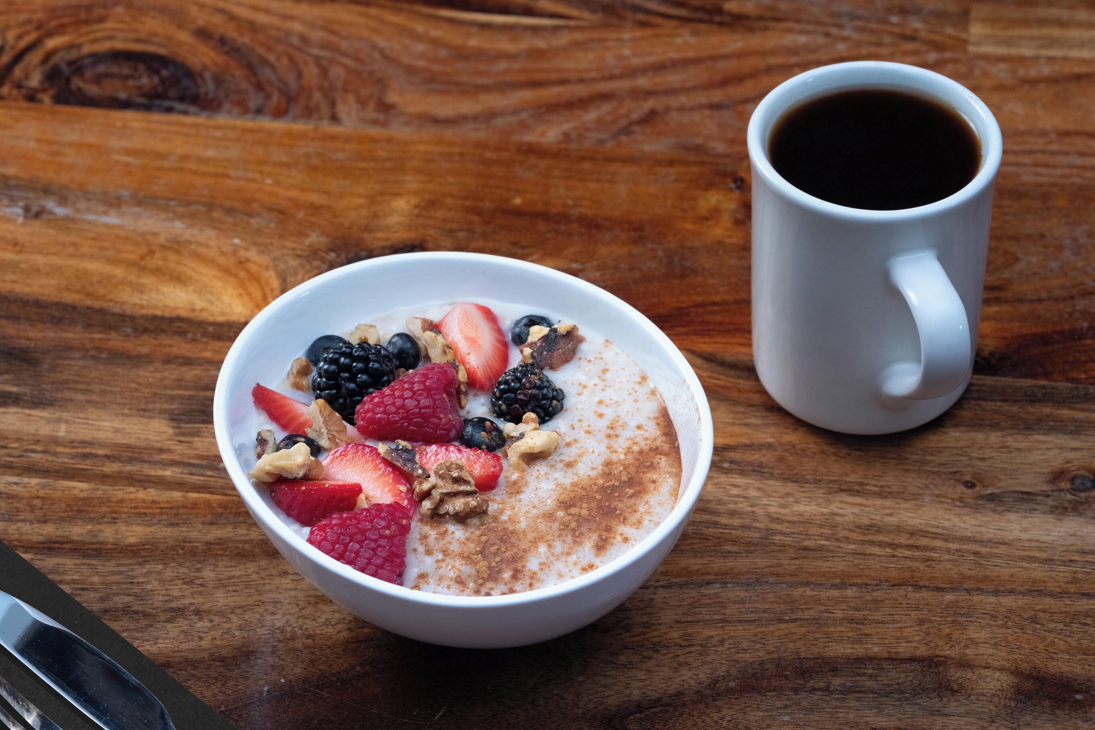 Un tazón de cereal con frutas y nueces, una taza blanca y una mesa de madera.