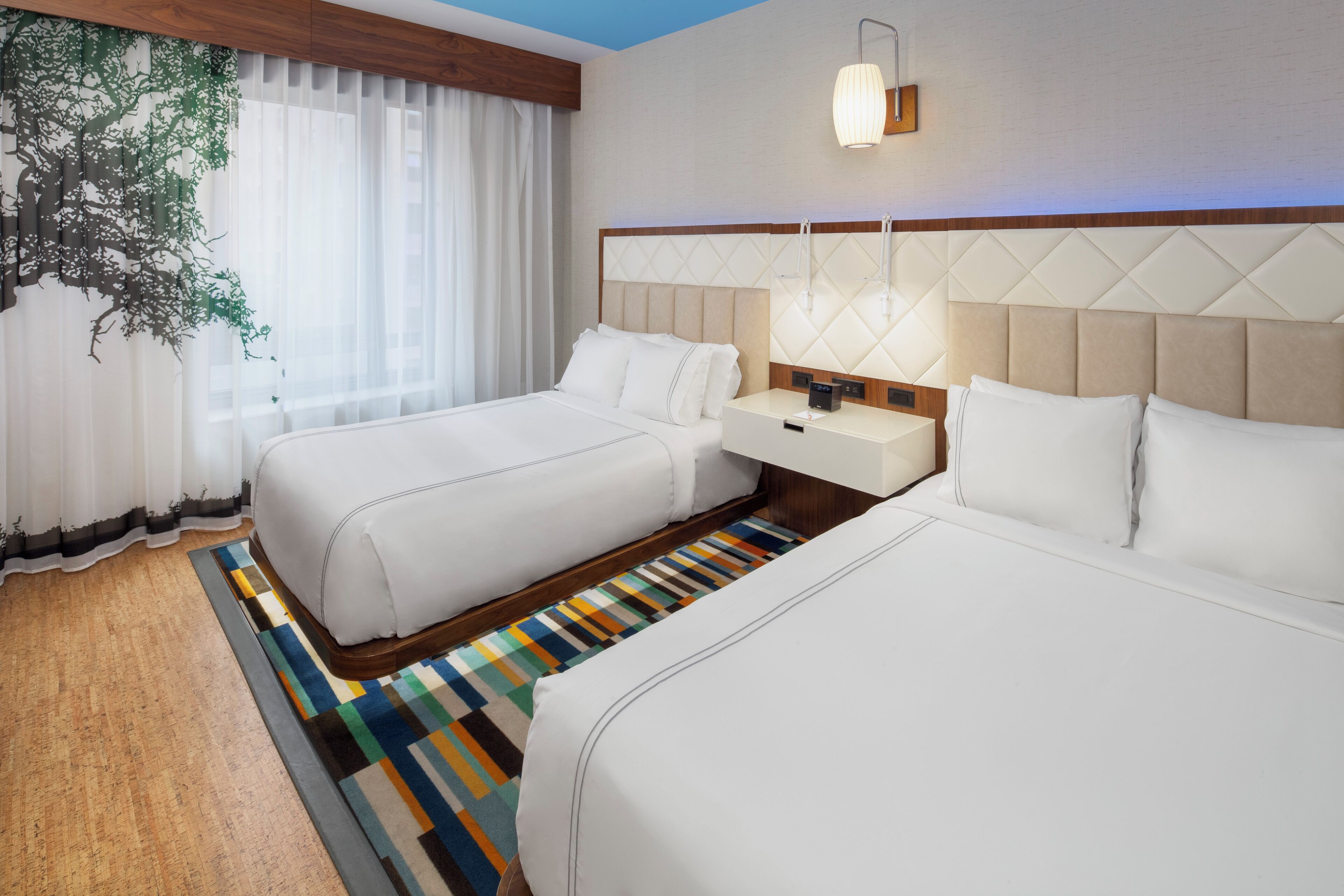 Habitación de hotel con dos camas, una mesita de noche, una lámpara, una ventana con cortinas translúcidas y piso de madera.