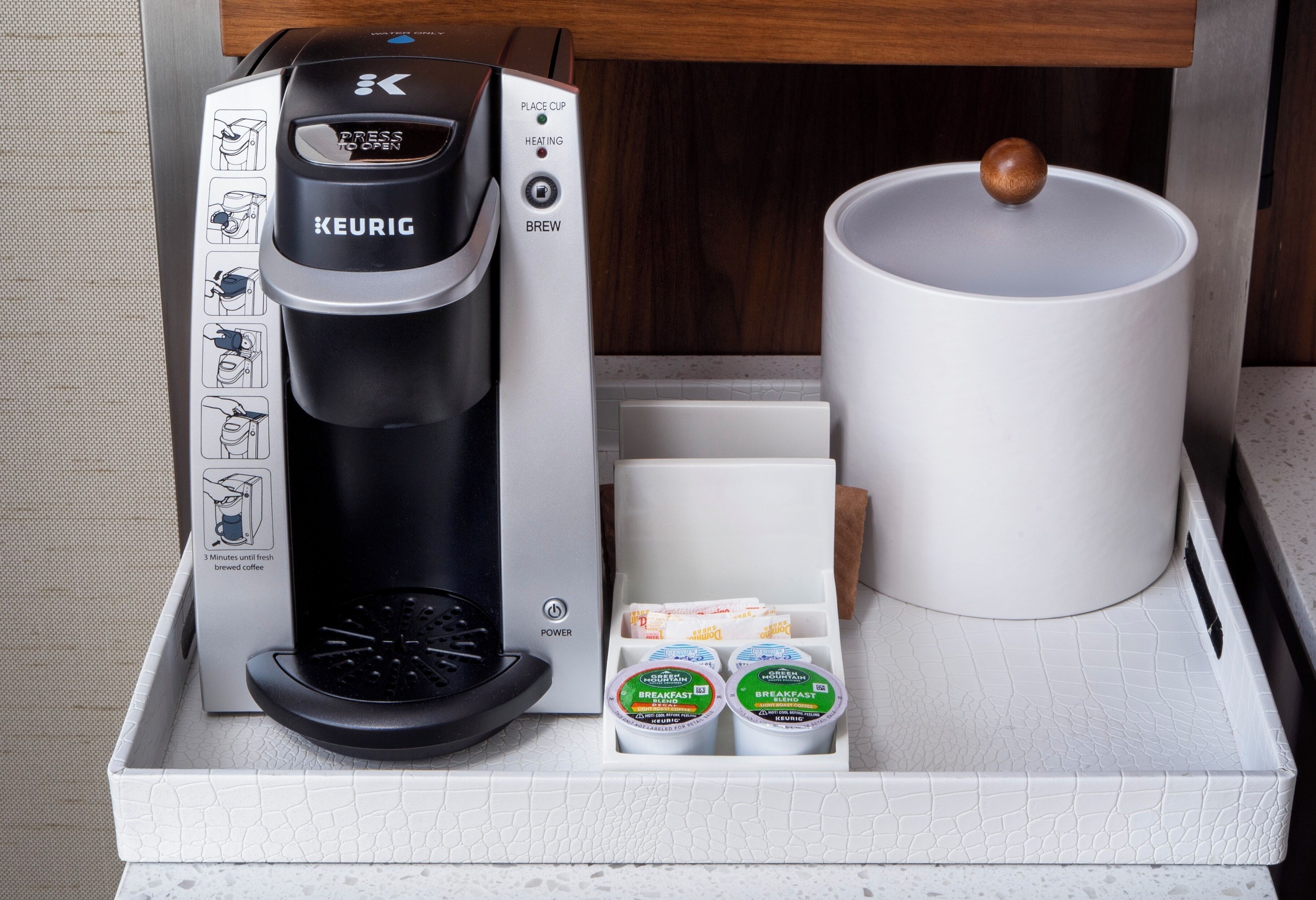 Una cafetera Keurig con un portacapsulas, una taza blanca y una bandeja con una cuchara de madera.