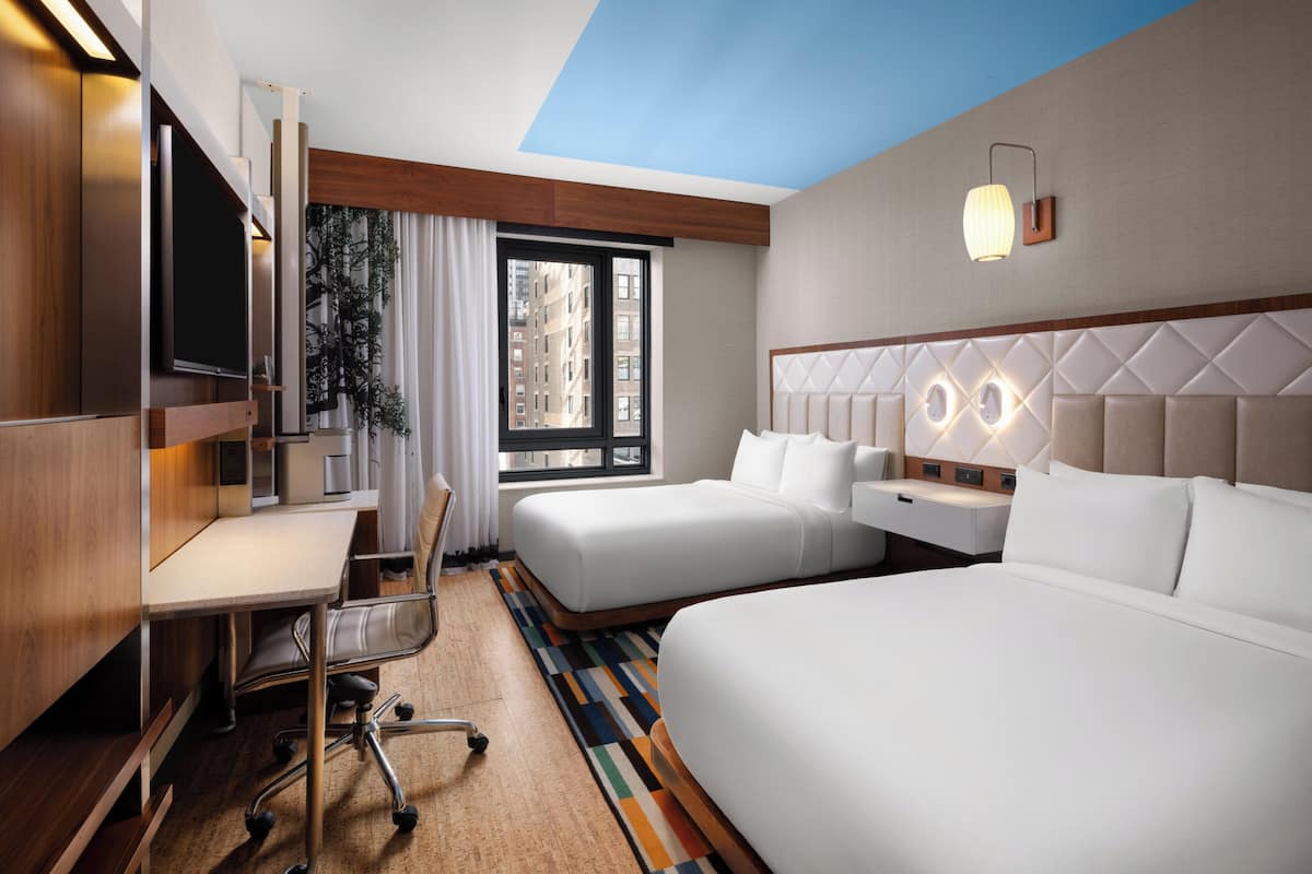Une chambre d’hôtel équipée d’un bureau, de deux lits, d’une télévision et d’une fenêtre donnant sur des immeubles.
