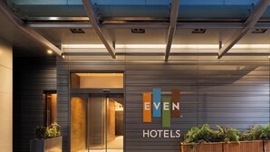 Een moderne hotelentree met een glazen deur, een bord waarop 'EVEN HOTELS' staat en een plantenbak met groen.