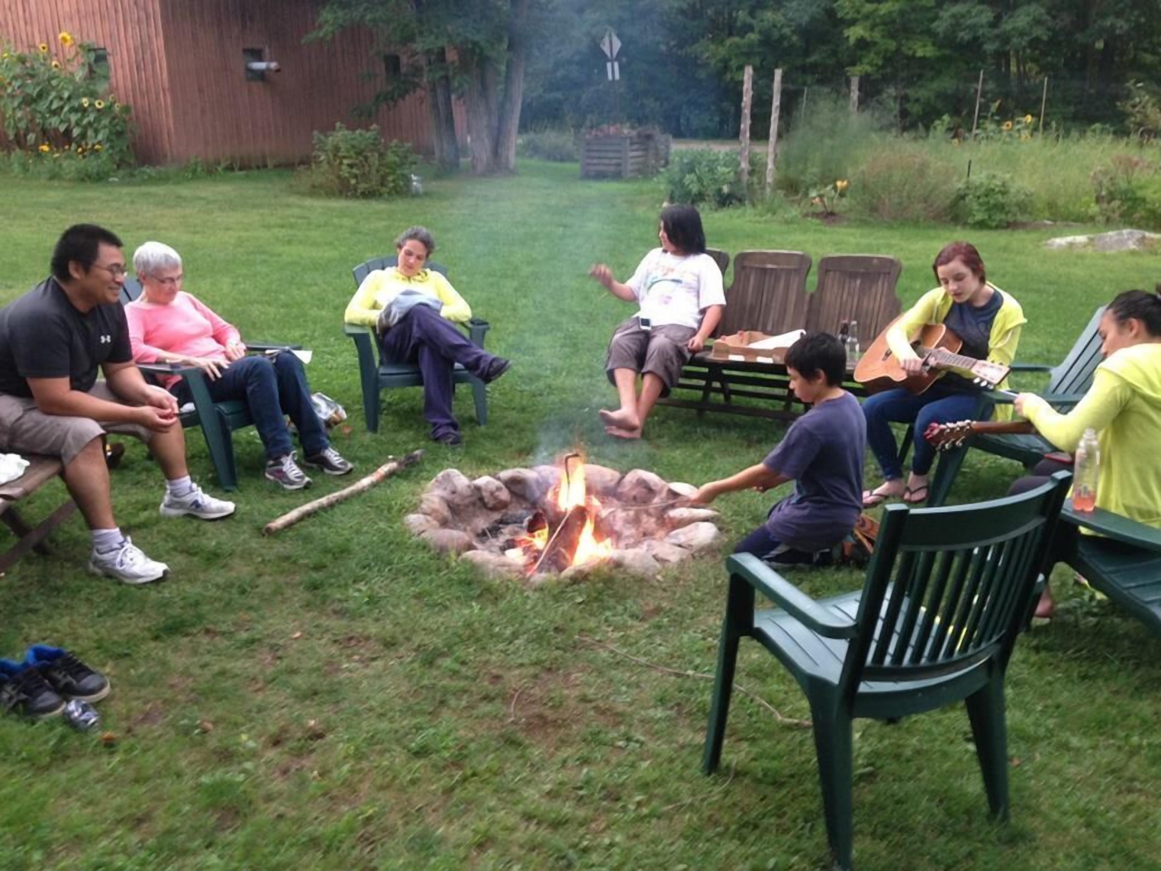 A group of people sitting around a campfire in a grassy outdoor area with chairs and a wooden fence in the background.