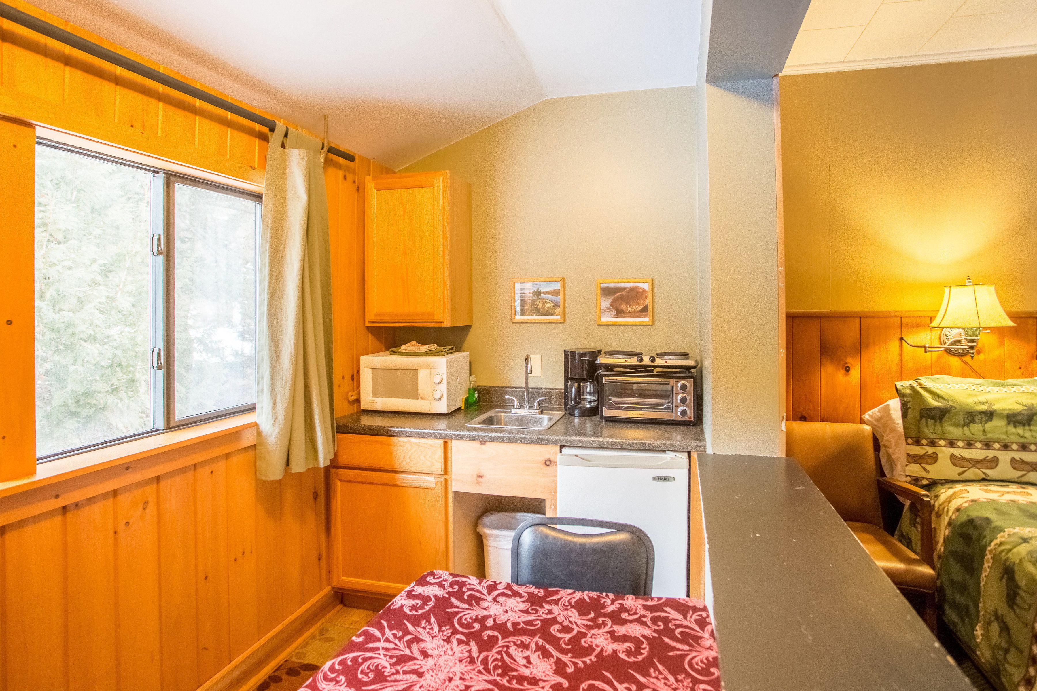 A compact kitchen with wooden cabinets, a microwave, a toaster, and a sink. There is a small dining table and a bed with patterned bedding.